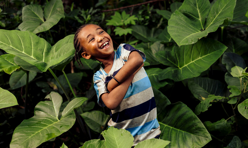 Ett etiopiskt barn poserar skrattande med armarna i kors mitt bland en stor buskes blad.