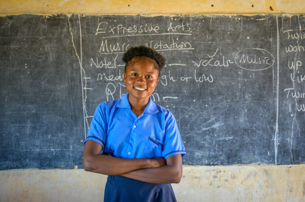 En flicka står leende med armarna i kort framför en skoltavla.