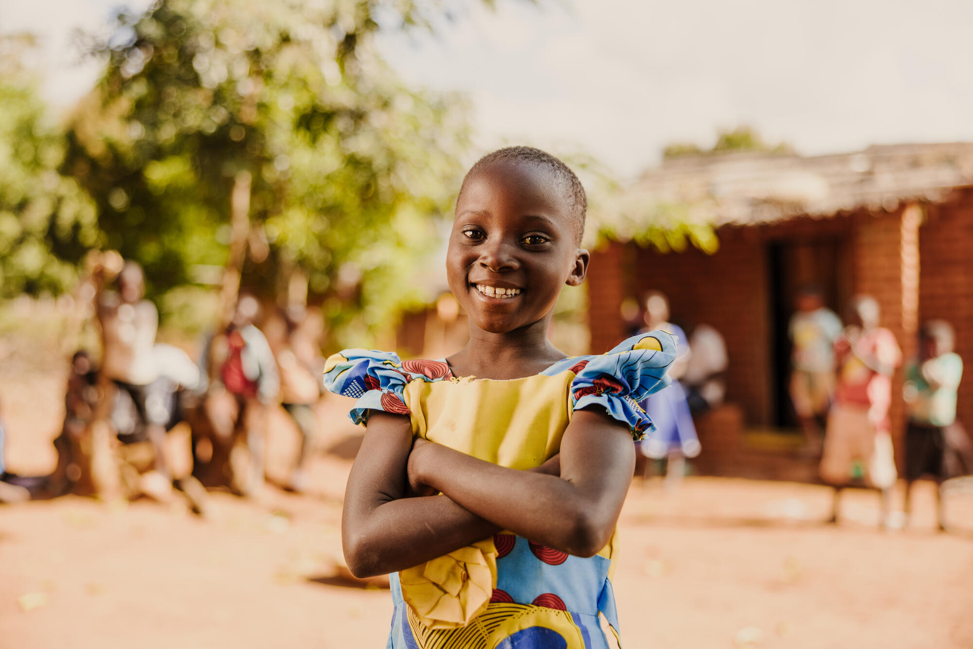 En flicka i Malawi, iklädd en blågul klänning, ler mot kameran med armarna i kors över bröstet.