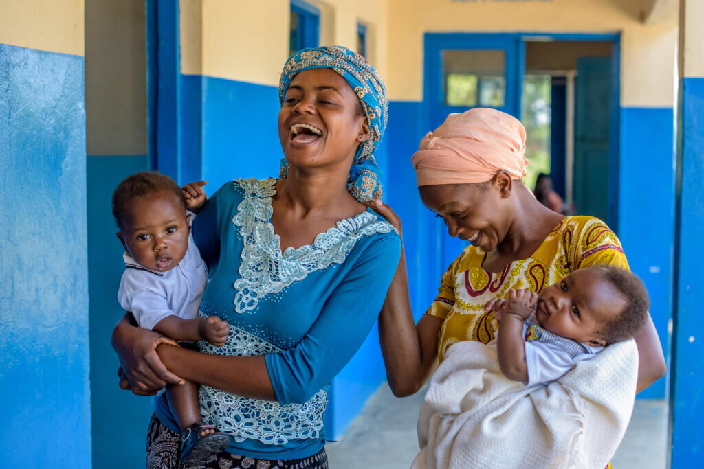 Två mammor i DRC skrattar tillsammans med sina barn i famnen.