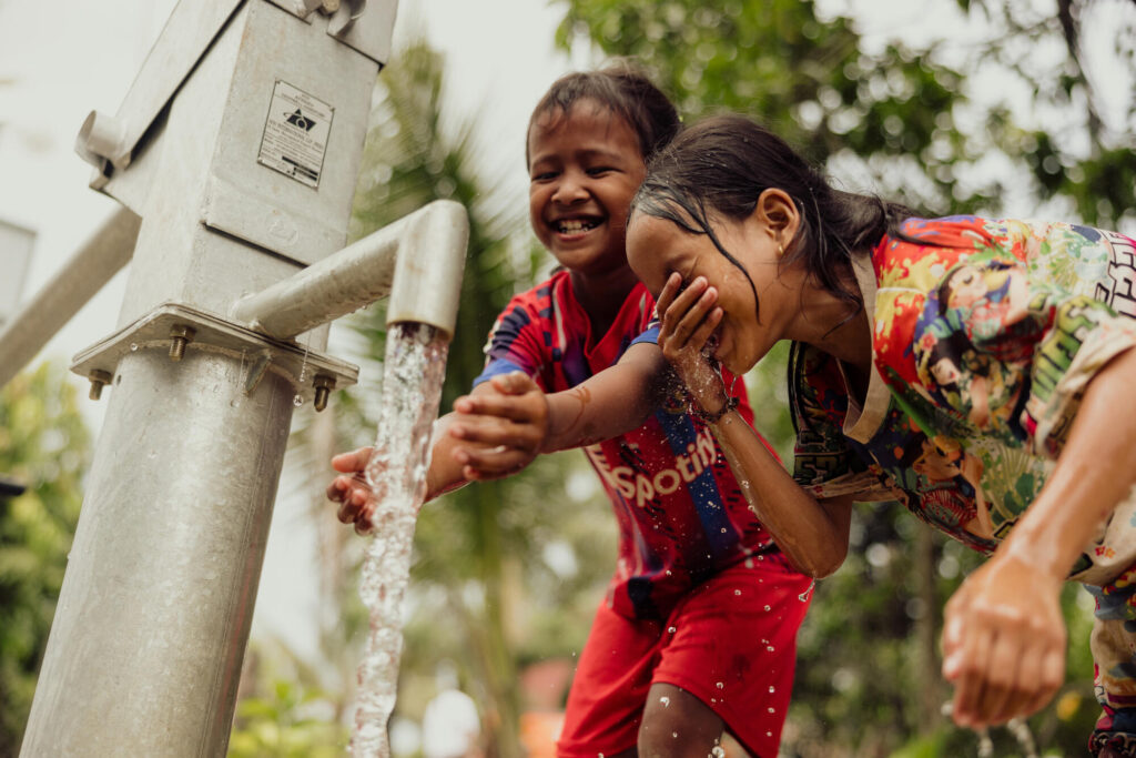 Två cambodjanska barn tvättar skrattande sina ansikten och händer vi en kran.