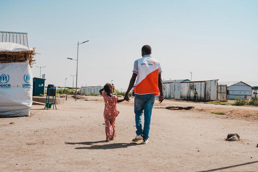 En World Vision medarbetare och en liten flicka går hand i hand genom ett flyktingläger i Södra Sudan.