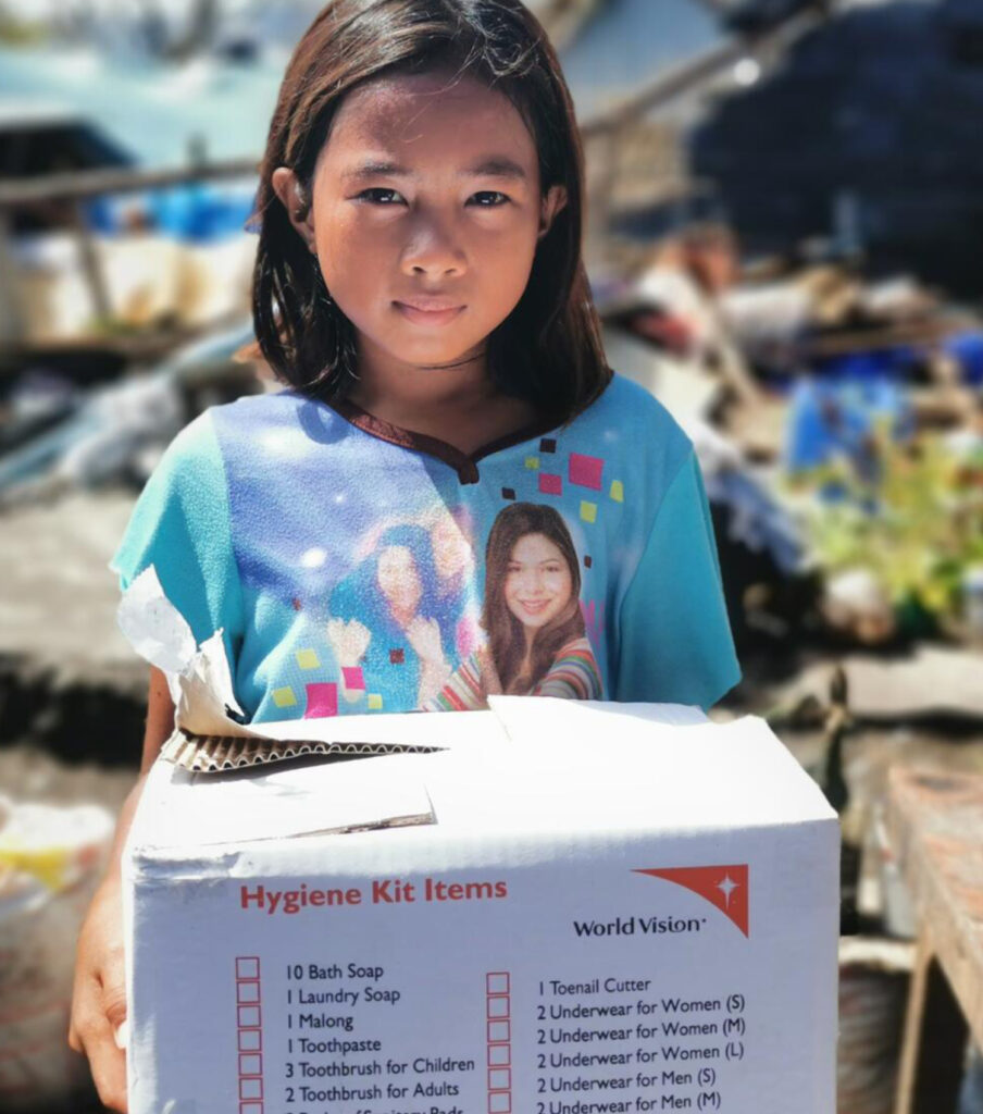 En flicka ser allvarligt in i kameran med ett nödhjälpspaket i famnen.