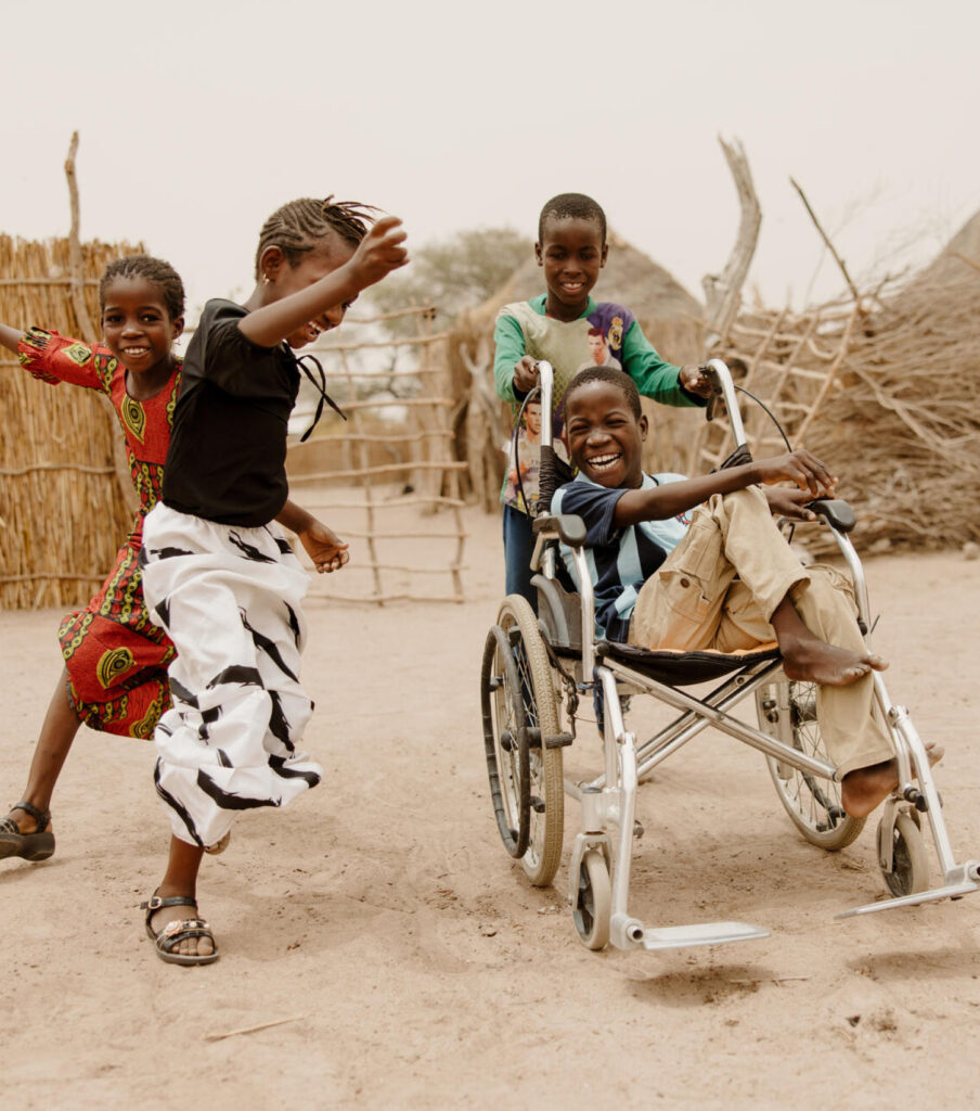 Fyra barn i Senegal dansar skrattande tillsammans. Ett av barnen, en pojke, sitter i en rullstor som körs av ett av de andra barnen.