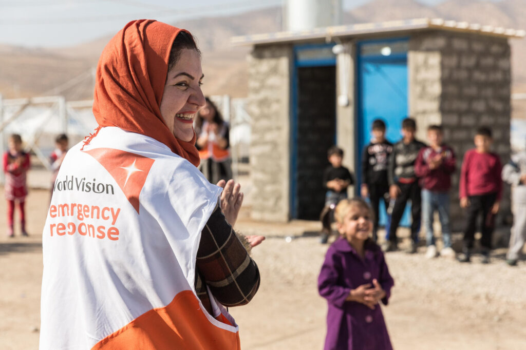 En irakisk World Vision volontär iklädd en World Vision väst med "Emergency response" på ryggen skrattar tillsammans med några barn.