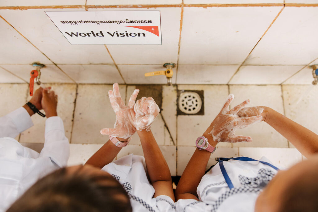 Närbild uppifrån av tre kambodjanska barns händer. Barnen tvättar händerna, som är full av tvållödder, vid ett handfat i kakel med varsin kran. Handfatet har en skylt med World Visions logo.