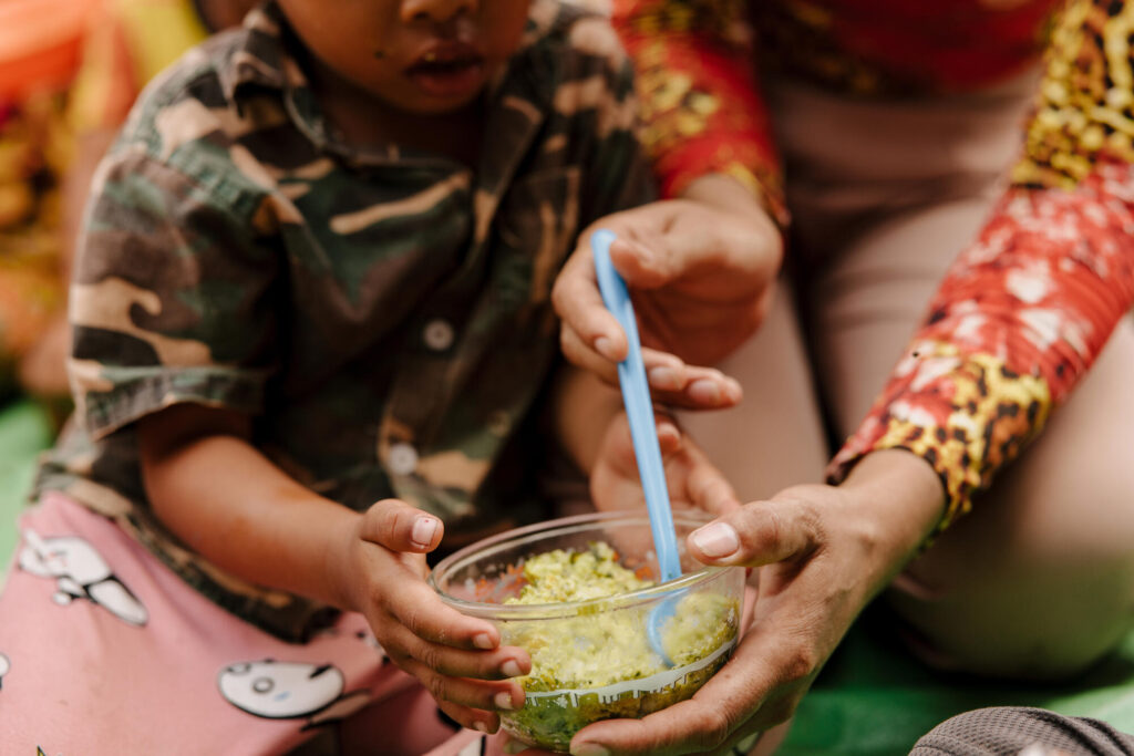 Närbild på en skål med mat. Ett par vuxna händer håller i skålen och en sked. Ett barn sitter bredvid och äter av maten samtidigt som även hen håller i skålen.