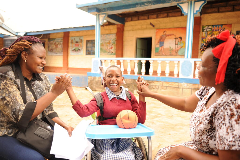 En kenyansk skolflicka i rullstol skrattar tillsammans med två vuxna kvinnor.