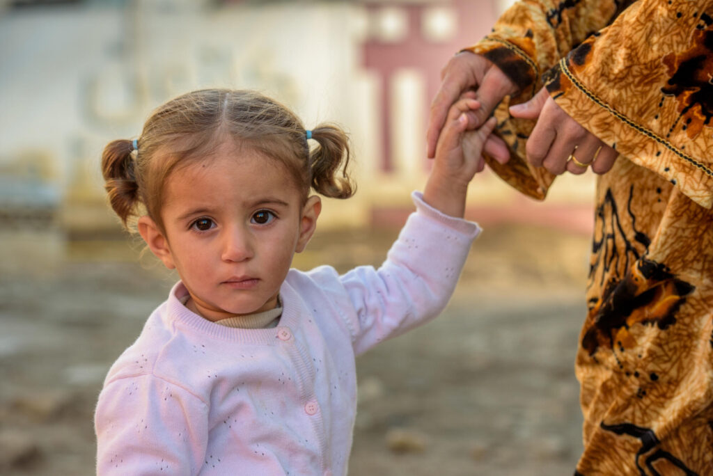En liten libanesisk flicka ser allvarligt in i kameran medan hon håller en kvinna i handen.