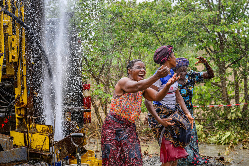 Tre kvinnor dansar skrattande i vattnet som sprutar upp ur marken då en borrmaskin som borrar brunnar äntligen funnit vatten.