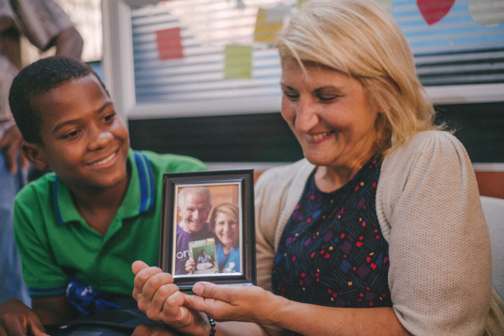 En World Vision fadder sitter tillsammans med sitt fadderbarn och håller i en inramad bild på sig själv och sin partner som håller i fadderbarnets bild. Både faddern och barnet ler tillsammans.