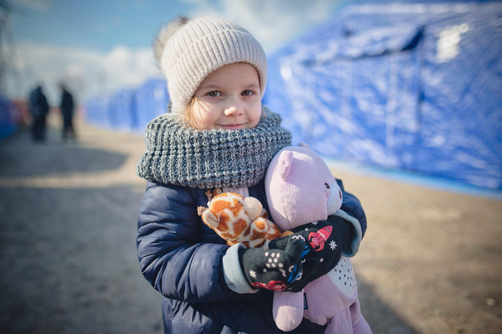 Ukrainska Lidya ler mot kameran med två gosedjur i armarna. Flickan är ca 5 år gammal. Lidya fotograferades iklädd vinterkläder vid gränsen till Rumänien kort efter att kriger startade i februari 2022.